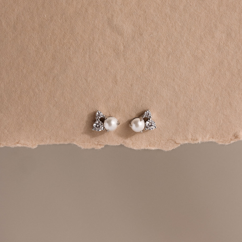The Pearl Diamond Cluster Studs in Sterling Silver, featuring pearl centers and sparkling stones, are displayed on a textured beige background.