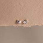 The Pearl Diamond Cluster Studs in Sterling Silver, featuring pearl centers and sparkling stones, are displayed on a textured beige background.