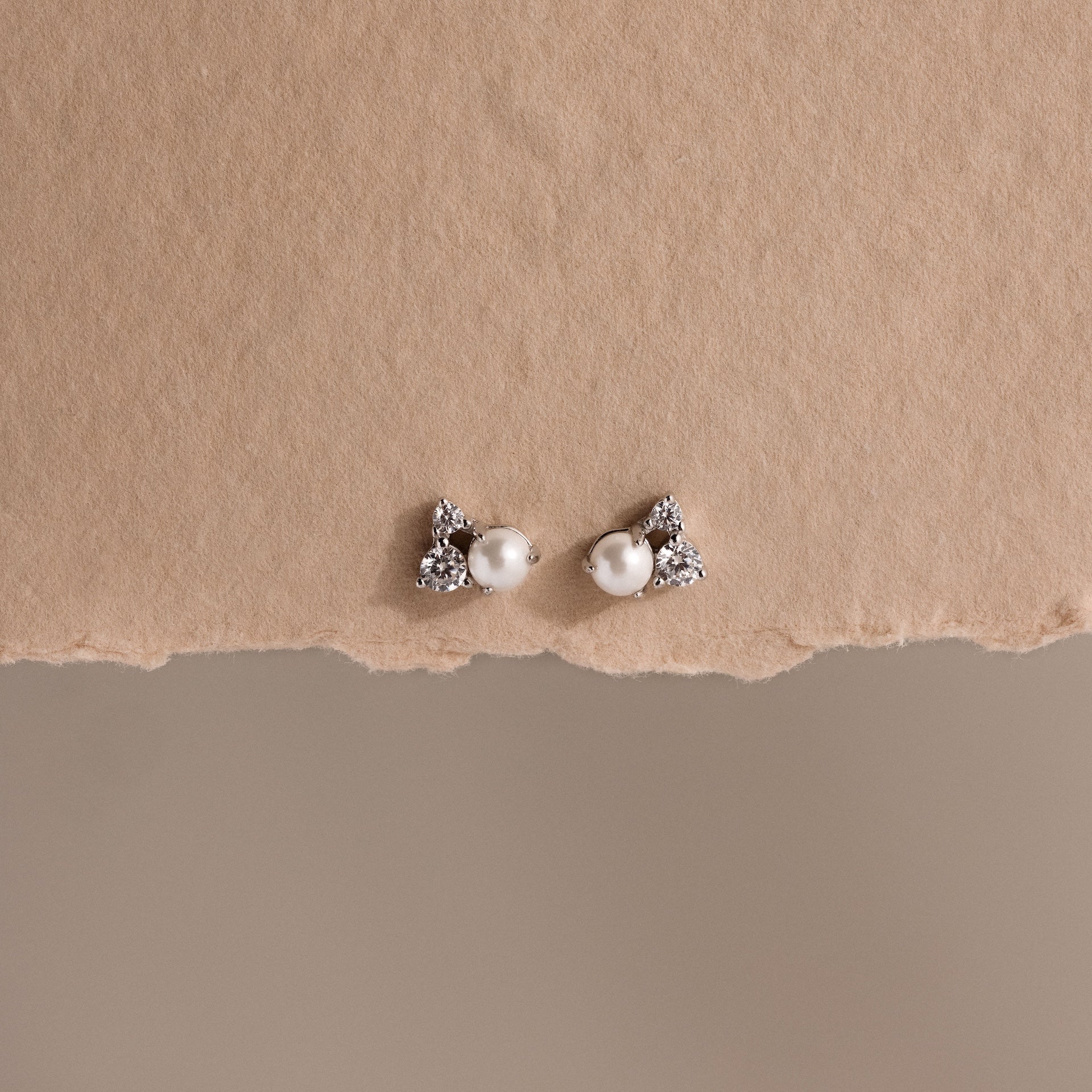 The Pearl Diamond Cluster Studs in Sterling Silver, featuring pearl centers and sparkling stones, are displayed on a textured beige background.