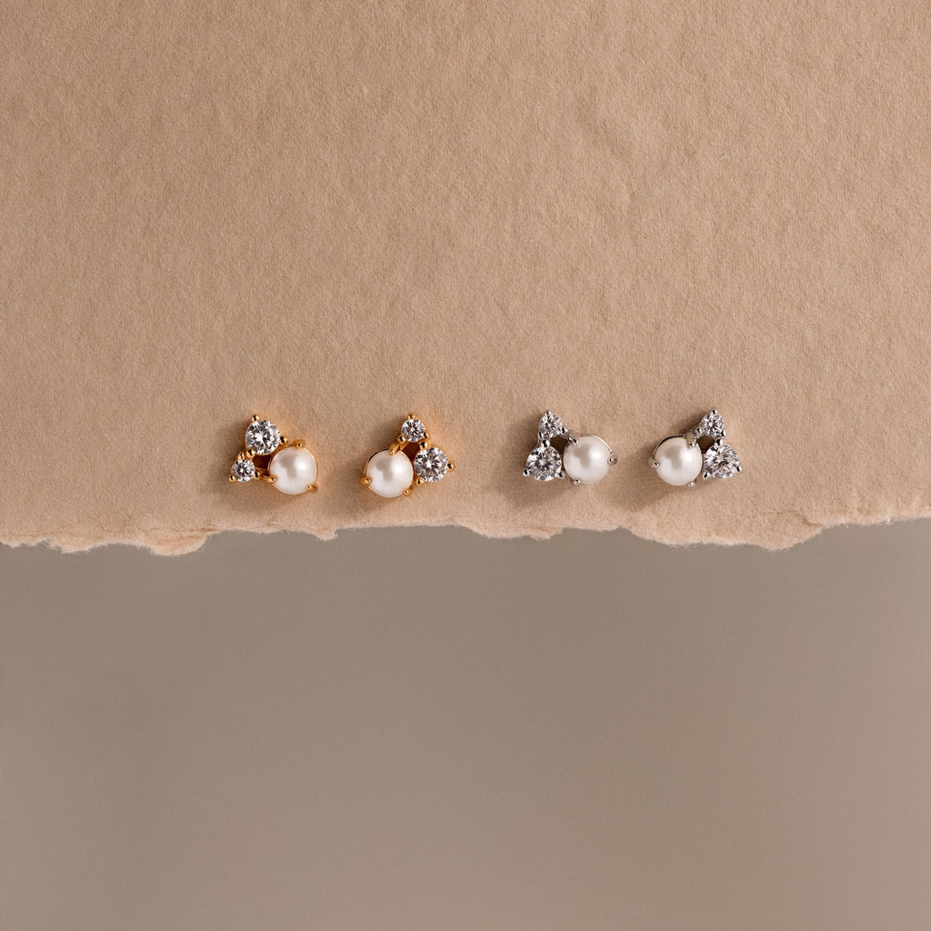 Two pairs of Pearl Diamond Cluster Studs, featuring pearls, CZ diamond accents, and crystals—one in gold and one in silver—are displayed on a textured surface.
