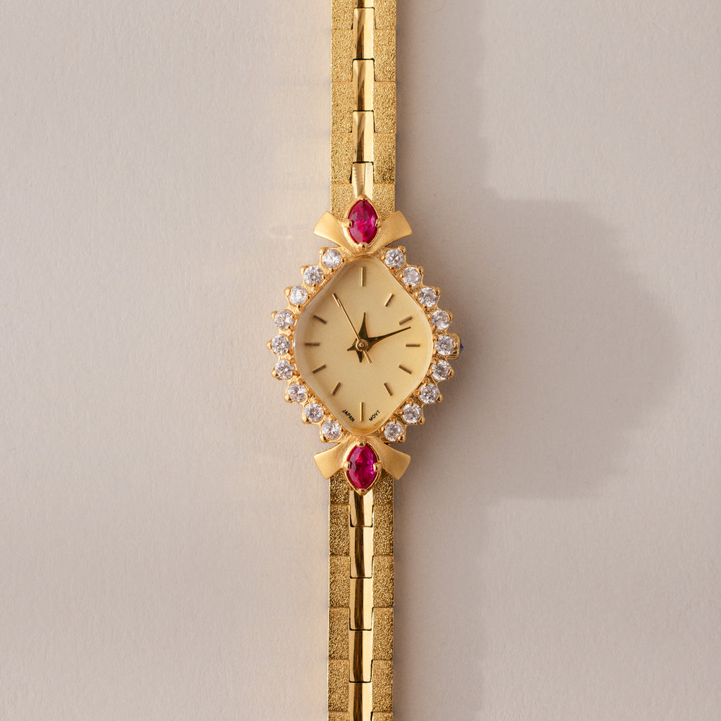 The Art Deco Ruby Timepiece features a gold bracelet and a diamond and ruby-studded face, radiating retro glamour and timeless elegance—set against a light beige background.