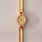 The Art Deco Ruby Timepiece features a gold bracelet and a diamond and ruby-studded face, radiating retro glamour and timeless elegance—set against a light beige background.