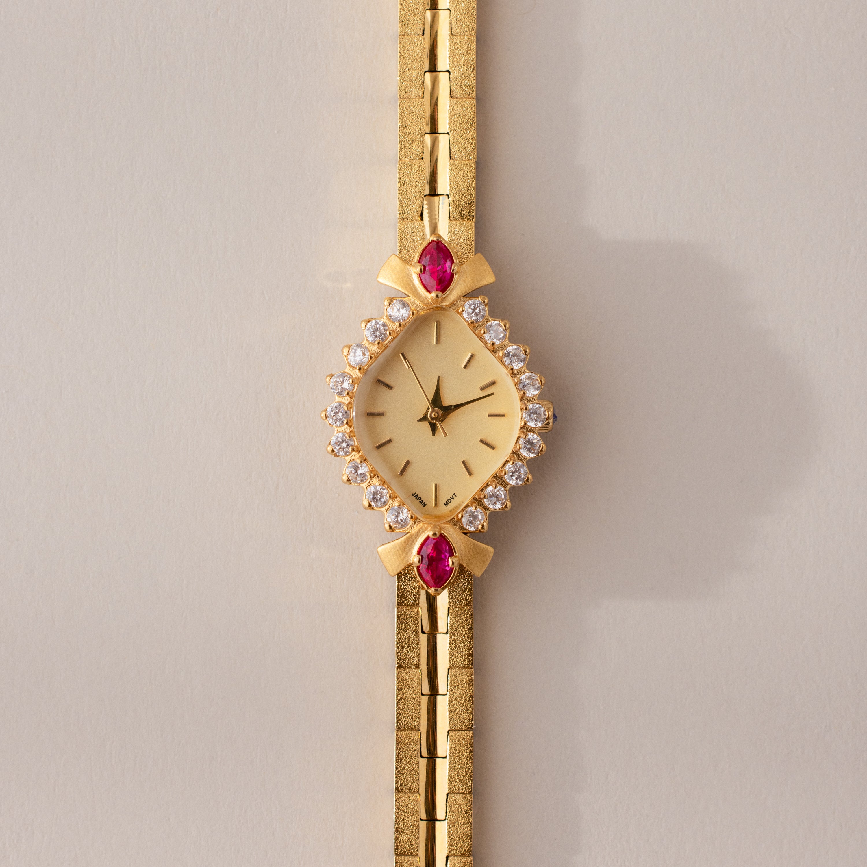 The Art Deco Ruby Timepiece features a gold bracelet and a diamond and ruby-studded face, radiating retro glamour and timeless elegance—set against a light beige background.