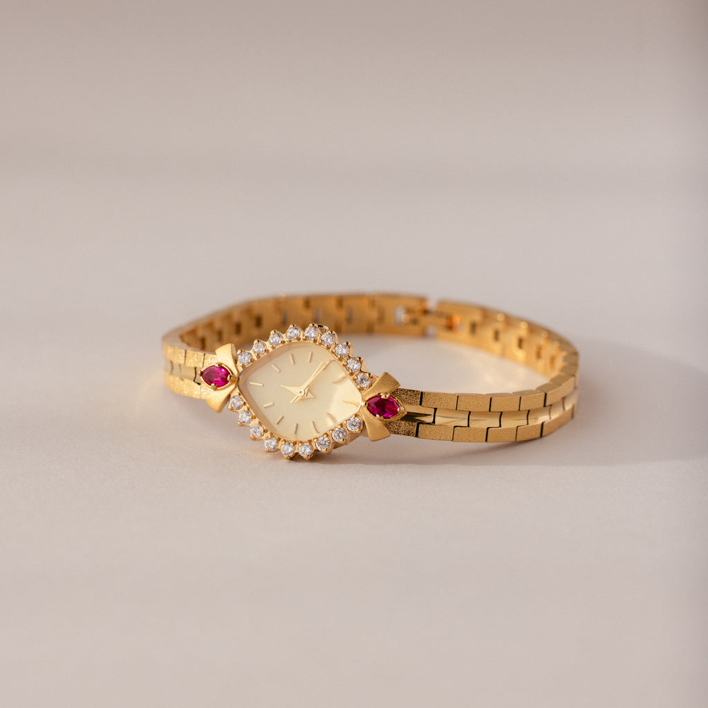 The Art Deco Ruby Timepiece features a gold wristwatch with a diamond-studded face and red gemstone accents, capturing retro glamour against a light background.