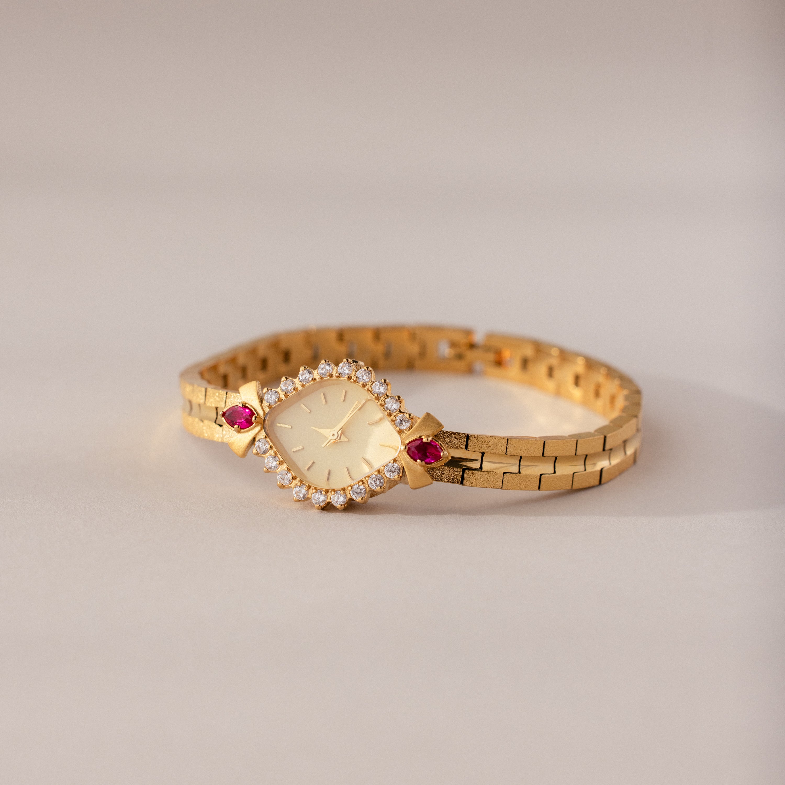 The Art Deco Ruby Timepiece features a gold wristwatch with a diamond-studded face and red gemstone accents, capturing retro glamour against a light background.