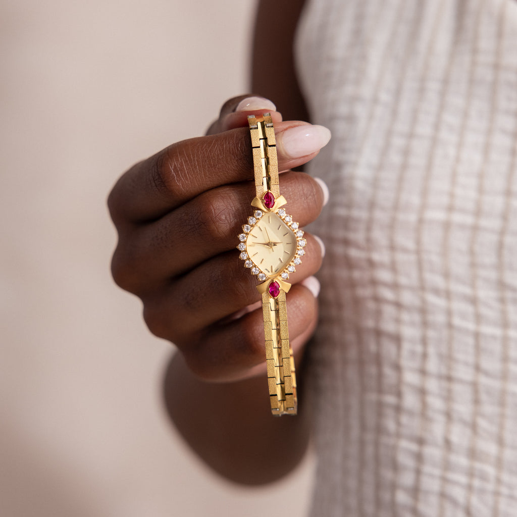 A hand holds the Art Deco Ruby Timepiece, a gold bracelet watch featuring red and white gemstones, shining with retro glamour against a textured white top.