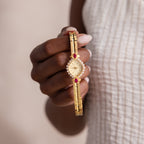A hand holds the Art Deco Ruby Timepiece, a gold bracelet watch featuring red and white gemstones, shining with retro glamour against a textured white top.