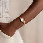 The Art Deco Ruby Timepiece, a gold bracelet wristwatch with a decorative face, channels retro glamour on your wrist when paired with cream-colored pants and a hand in pocket.
