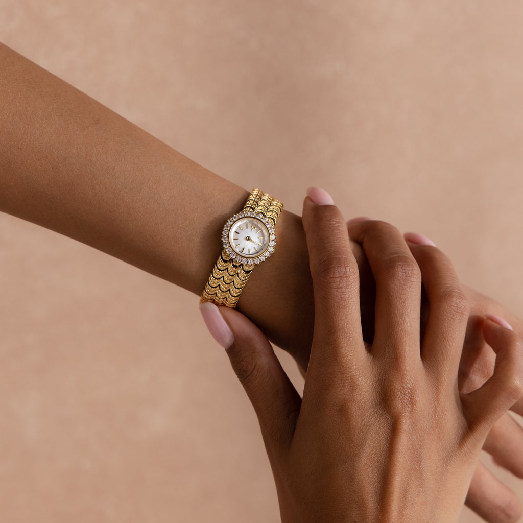 A hand adjusts the Pavé Scalloped Timepiece in 18K Gold, featuring a round jeweled face and pavé bezel, on another wrist against a beige background.