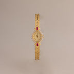 The Art Deco Ruby Timepiece features a gold bracelet and gemstone accents on the face, evoking retro glamour, shown against a plain beige background.