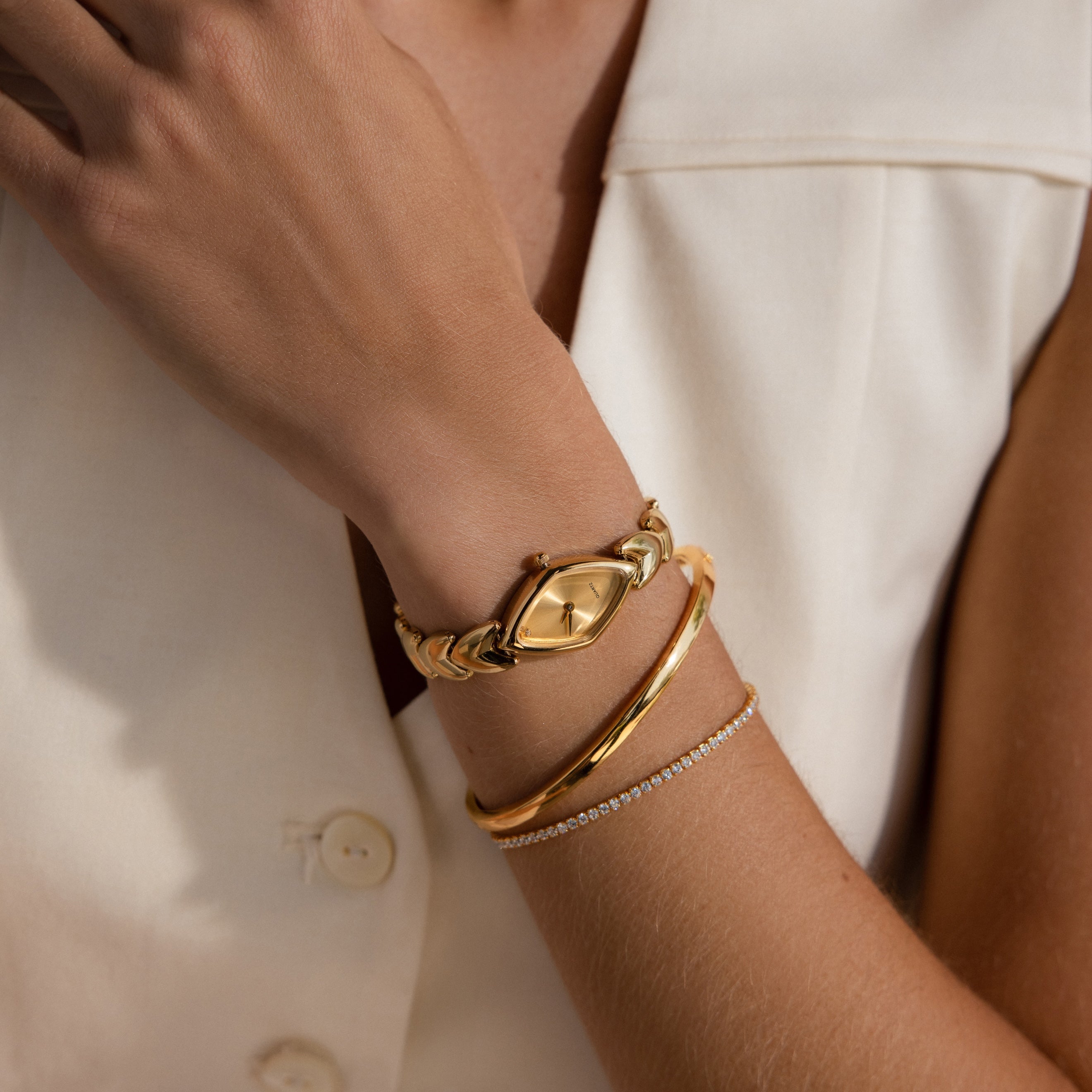 Close-up of a woman's wrist adorned with a gold oval-faced watch, paired with a gold bangle and a delicate diamond tennis bracelet, worn with a sleeveless cream blouse.