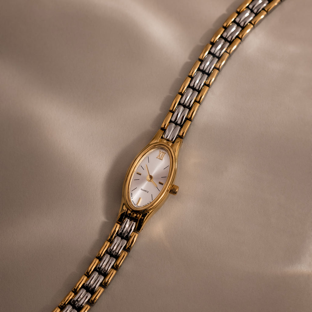 The Slim Oval Timepiece, featuring a gold and silver polished link bracelet, rests elegantly on a soft beige surface.