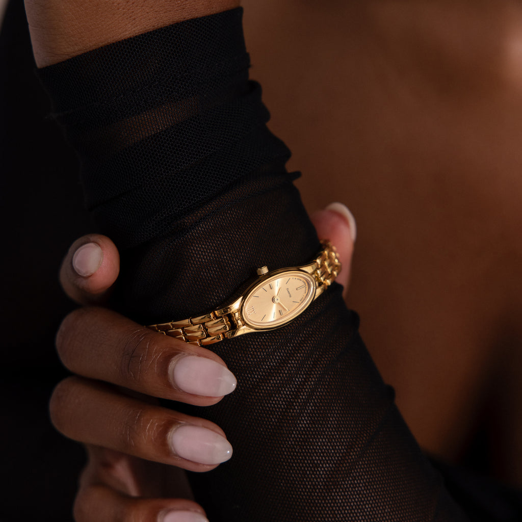 A hand in a sheer black glove gently holds the wrist, showcasing the Slim Oval Timepiece with a polished link bracelet.