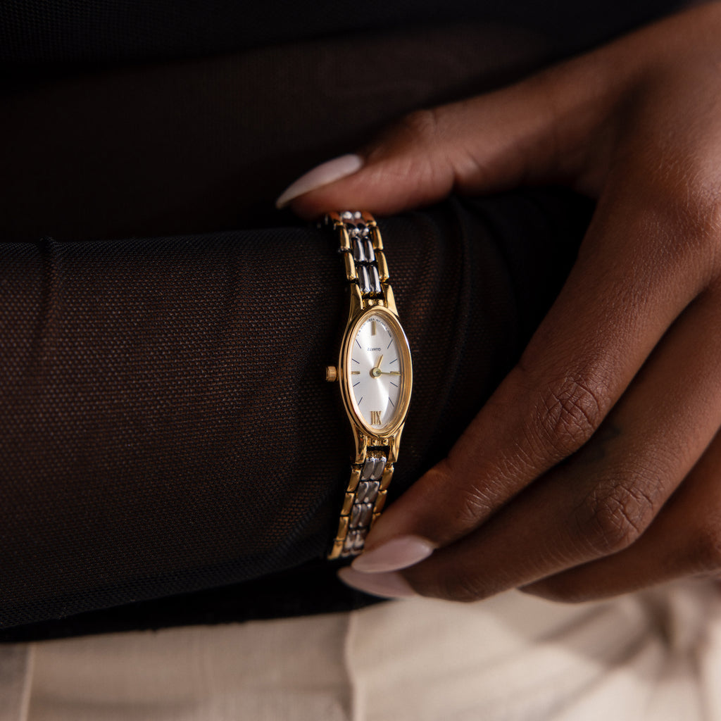 A hand showcases the Slim Oval Timepiece, featuring a polished link bracelet and an oval face, styled over a sheer black sleeve.