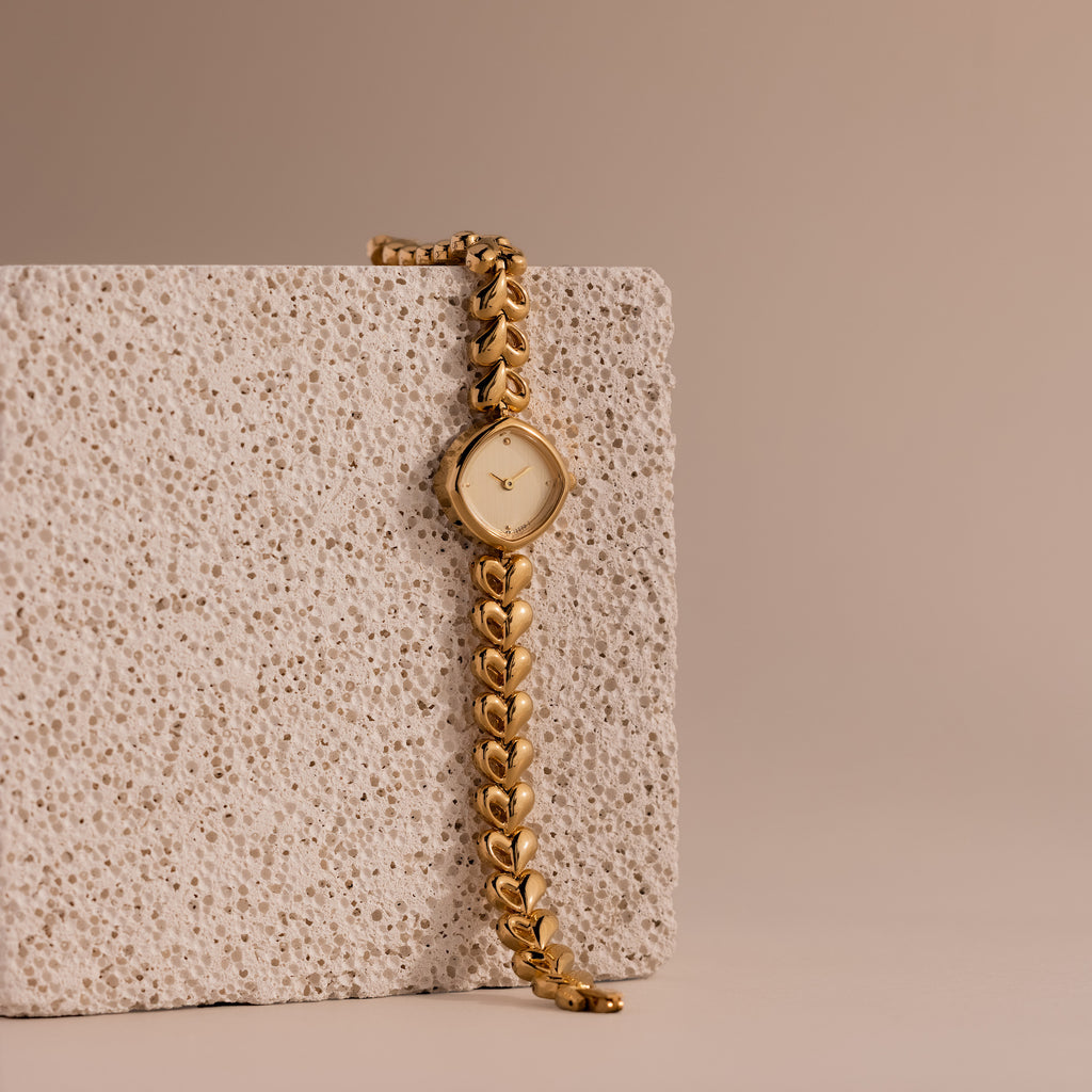 The Heart Link Timepiece, featuring heart-shaped links and a minimalist dial, drapes elegantly over a textured stone block against a beige background.