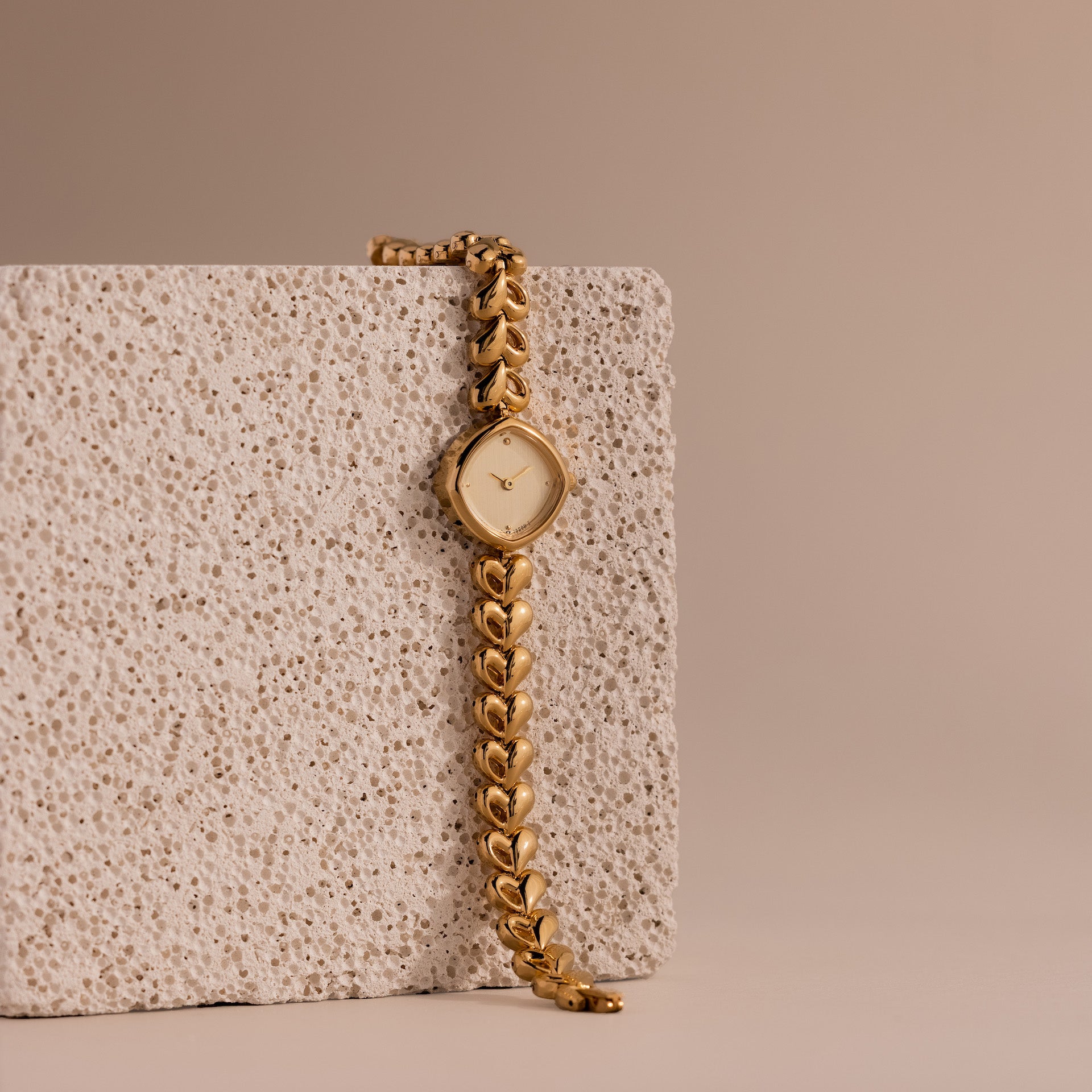 The Heart Link Timepiece, featuring heart-shaped links and a minimalist dial, drapes elegantly over a textured stone block against a beige background.