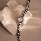 The Heart Link Timepiece, featuring a minimalist dial and heart-shaped links in silver, is displayed on a wavy, cream-colored surface.