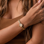 A woman wearing the Heart Link Timepiece, featuring heart-shaped links, crosses her arm over her chest while dressed in a brown ribbed top.