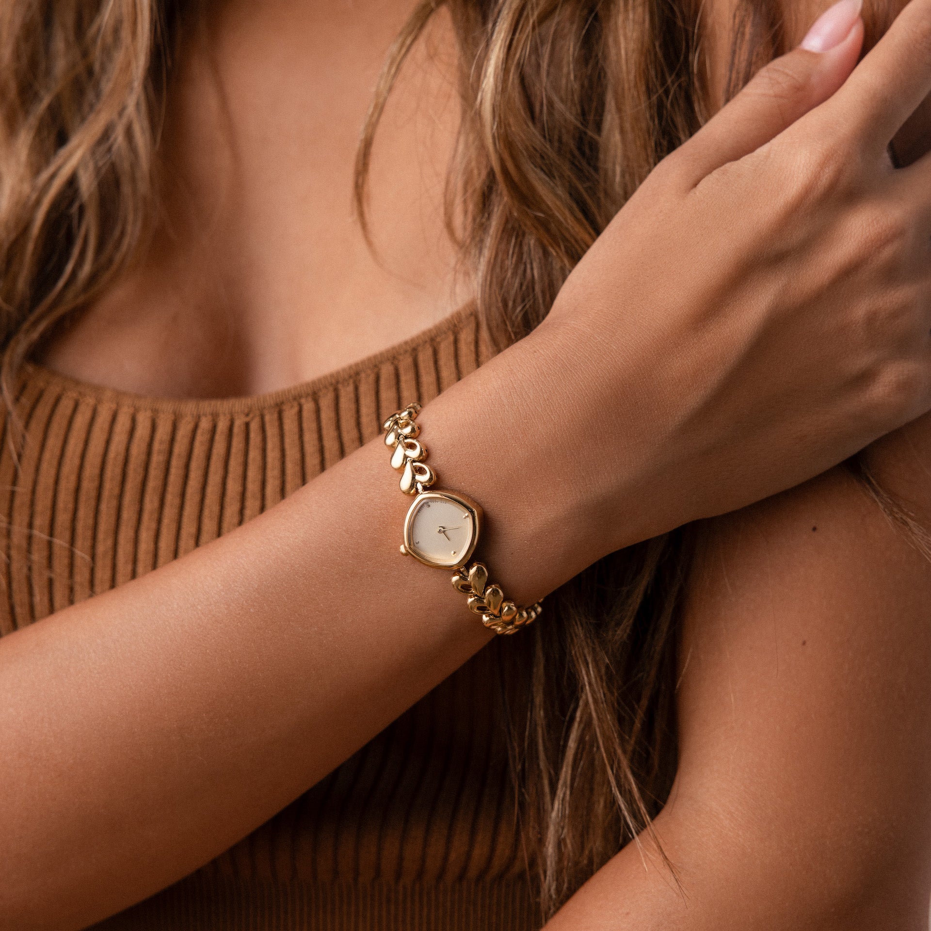 A woman wearing the Heart Link Timepiece, featuring heart-shaped links, crosses her arm over her chest while dressed in a brown ribbed top.