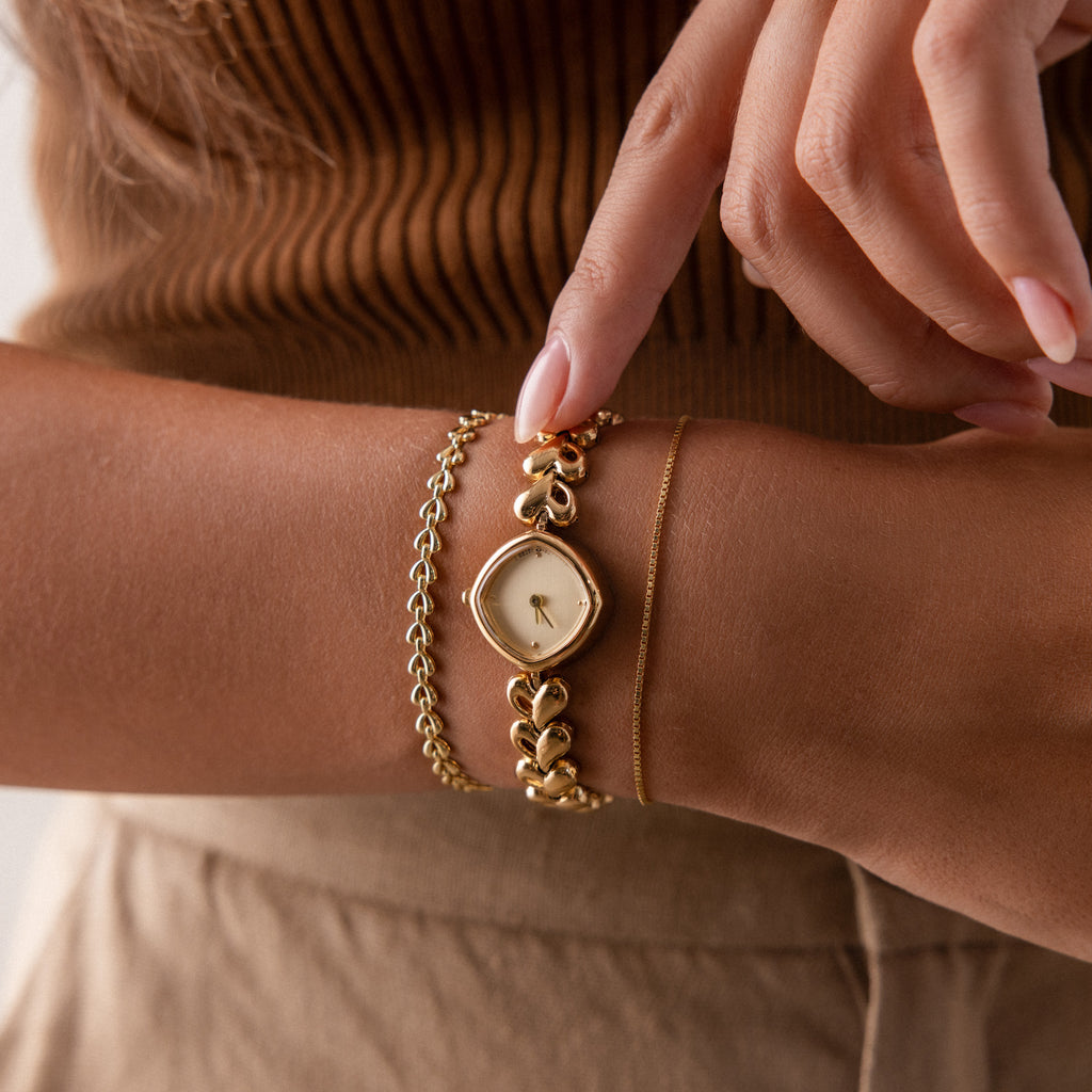 A woman wearing gold bracelets and the Heart Link Timepiece in 18K Gold, with heart-shaped links, gently touches the minimalist dial of her gold wristwatch.