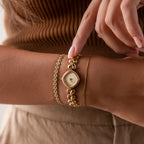 A woman wearing gold bracelets and the Heart Link Timepiece in 18K Gold, with heart-shaped links, gently touches the minimalist dial of her gold wristwatch.