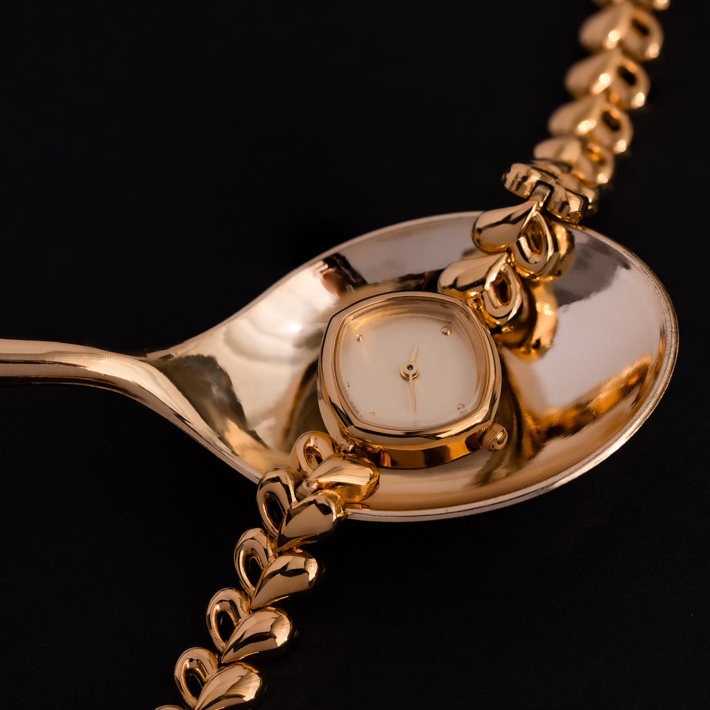 The Heart Link Timepiece in 18K Gold, featuring heart-shaped links and a minimalist dial, rests on a shiny gold spoon against a black background.