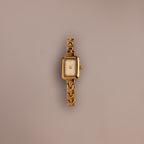 The Azteca Link Timepiece in 18K Gold features a rectangular case and minimalist champagne dial, with a patterned gold band, displayed elegantly on a plain light brown background.