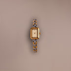 The Azteca Link Timepiece in Mixed Metal, featuring a rectangular case, patterned band, and minimalist champagne dial, rests on a plain beige background.