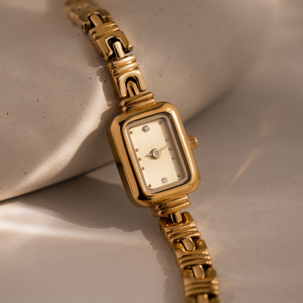 The Azteca Link Timepiece in 18K Gold, featuring a rectangular face and decorative band, rests on a smooth, light stone surface.