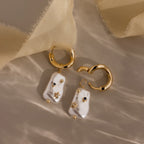 The Starry Baroque Pearl Charm Hoops in 18K Gold, featuring large irregular pearls and small star details, are displayed on a soft beige fabric background.