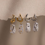 Two pairs of Starry Baroque Pearl Charm Hoops, featuring delicate pearl charms and gold hoops, displayed on a soft beige surface.