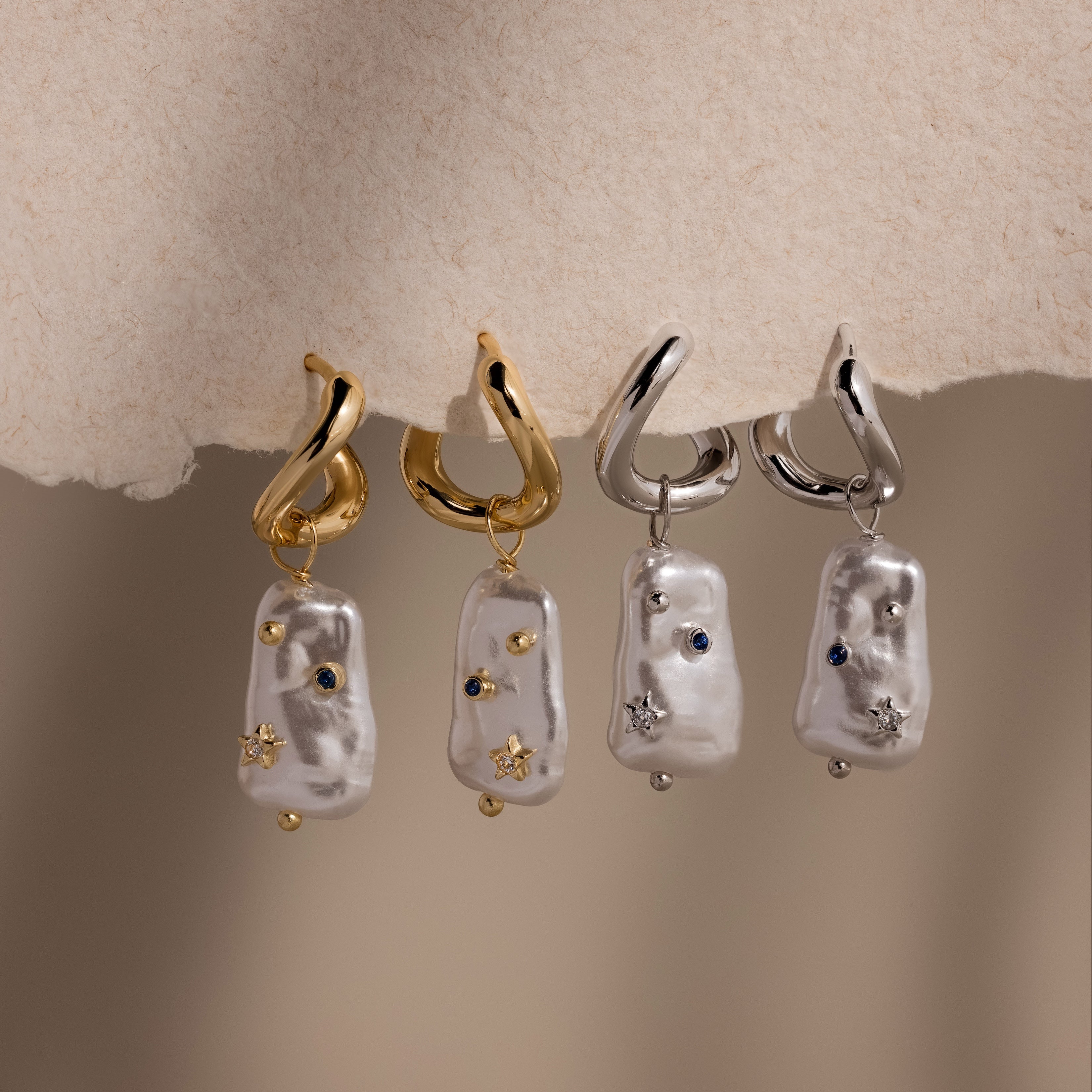 Two pairs of Starry Baroque Pearl Charm Hoops, featuring delicate pearl charms and gold hoops, displayed on a soft beige surface.