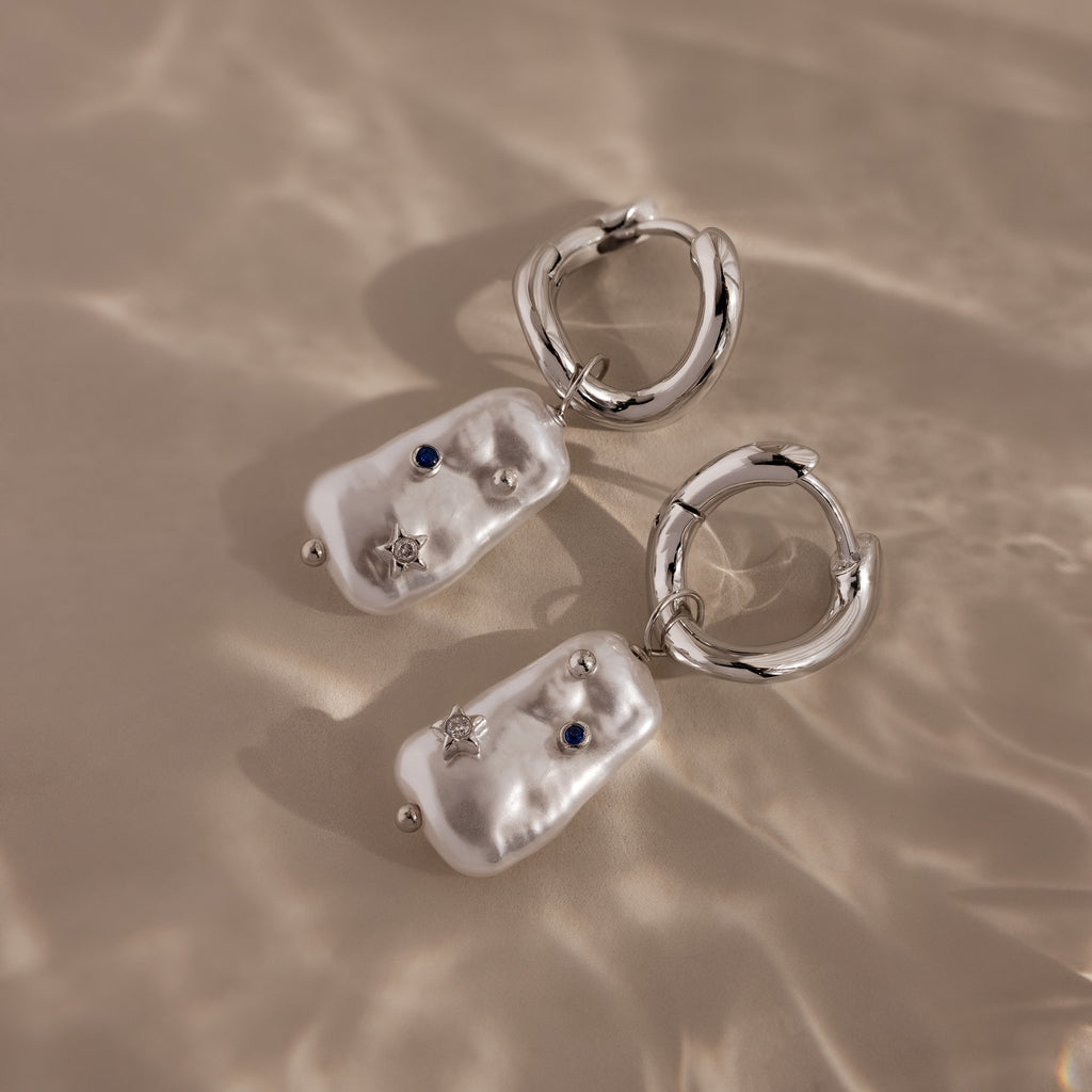 The Starry Baroque Pearl Charm Hoops in White Gold feature irregular pearl-like charms, blue stones, and star accents, capturing a unique allure reminiscent of Baroque pearl earrings, shown on a beige surface.