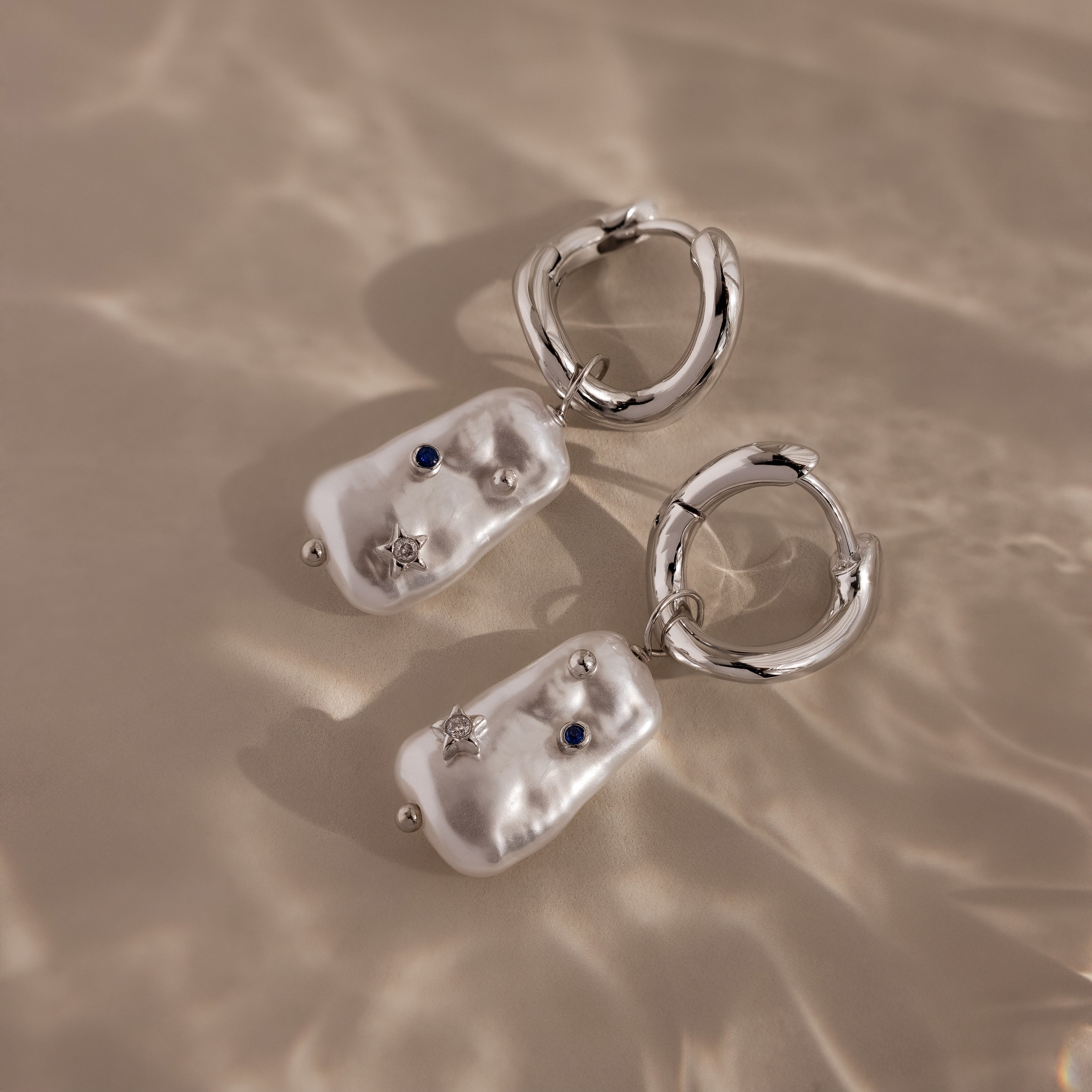 The Starry Baroque Pearl Charm Hoops in White Gold feature irregular pearl-like charms, blue stones, and star accents, capturing a unique allure reminiscent of Baroque pearl earrings, shown on a beige surface.