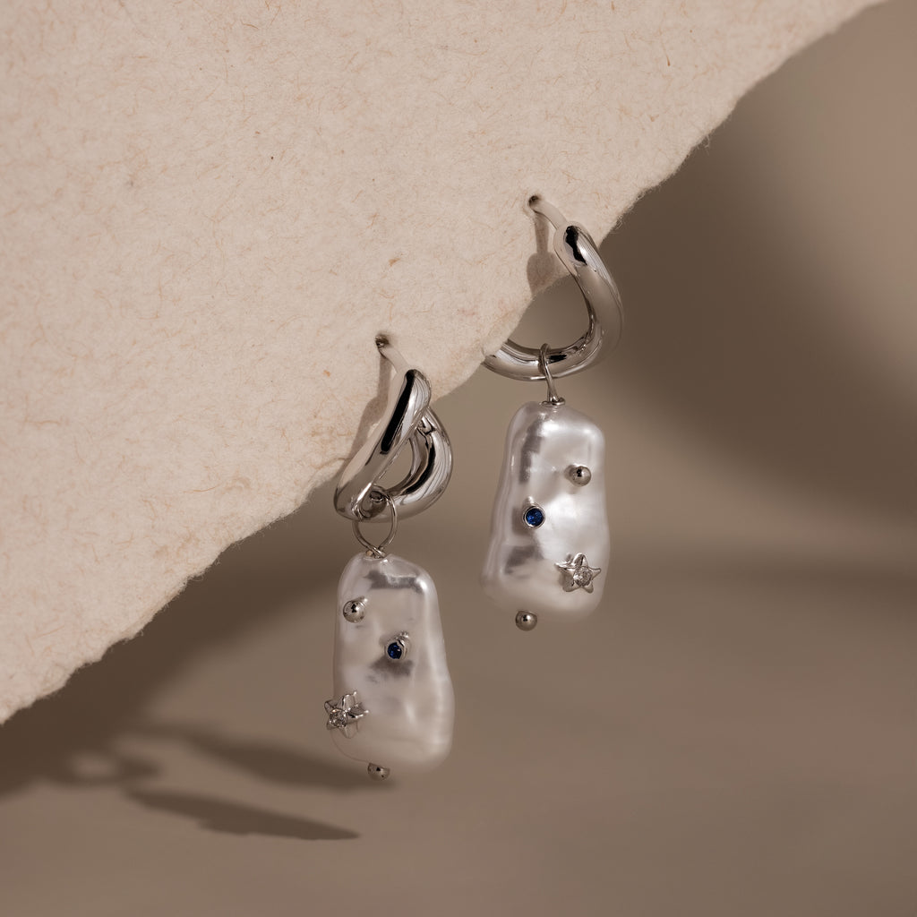 Starry Baroque Pearl Charm Hoops in White Gold feature irregular pearl drops with blue and silver accents, displayed on a textured beige surface.