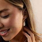 A woman smiles, touching her ear and showcasing the Starry Baroque Pearl Charm Hoops.