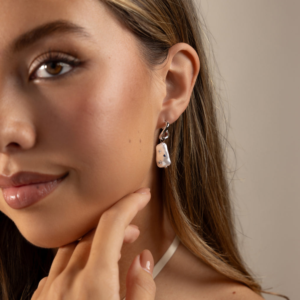 A woman with long hair wears the Starry Baroque Pearl Charm Hoops in White Gold, touching her chin and gazing slightly off-camera.