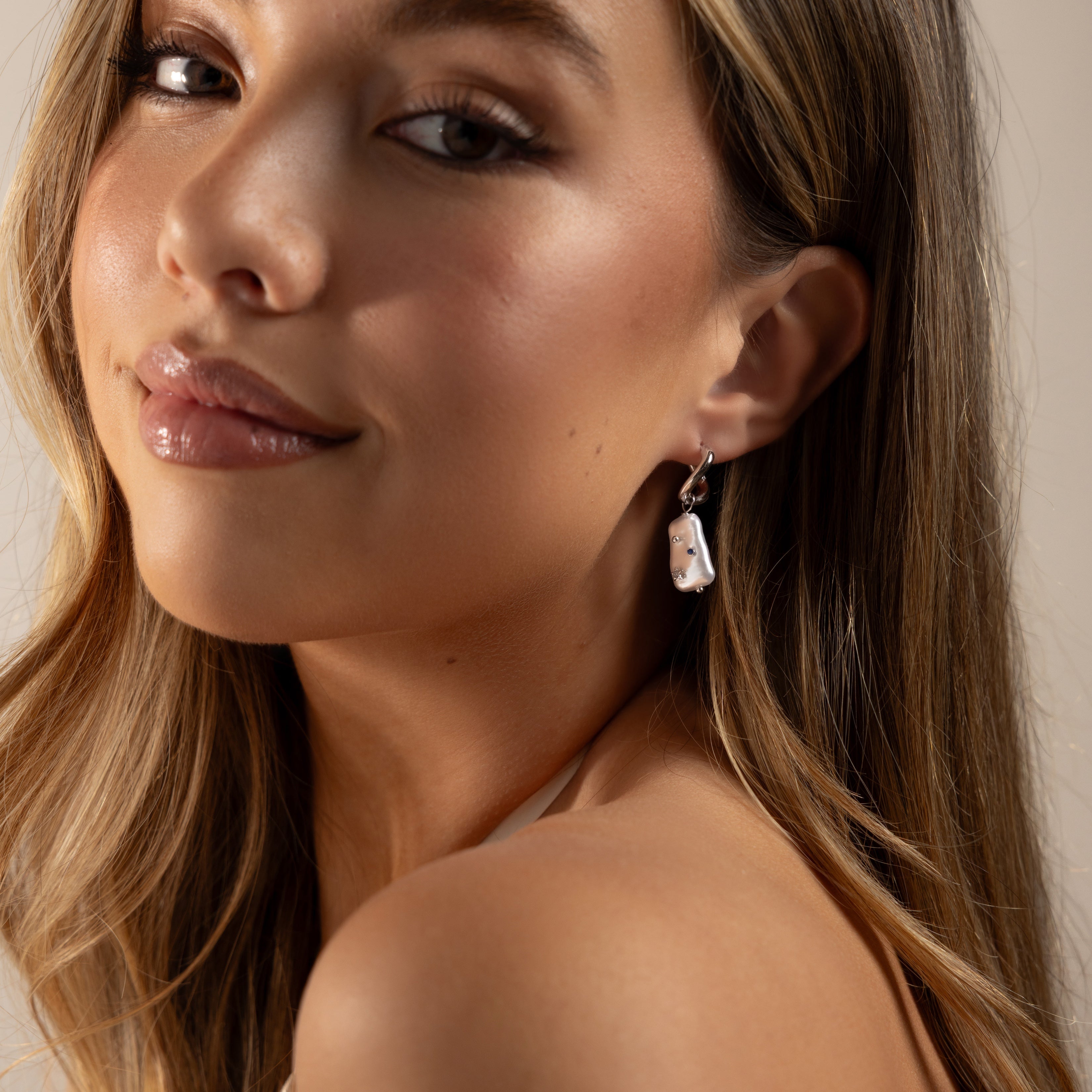A woman with long hair smiles softly, wearing natural makeup and elegant Starry Baroque Pearl Charm Hoops in White Gold.