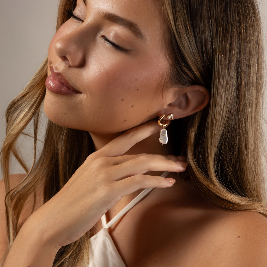 A woman with long hair and closed eyes touches her Starry Baroque Pearl Charm Hoops while wearing a white halter top.
