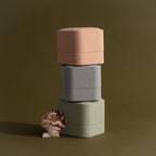 Three Suede Ring Boxes in pink, gray, and green are stacked beside a seashell on an olive background, highlighting their minimalist design.