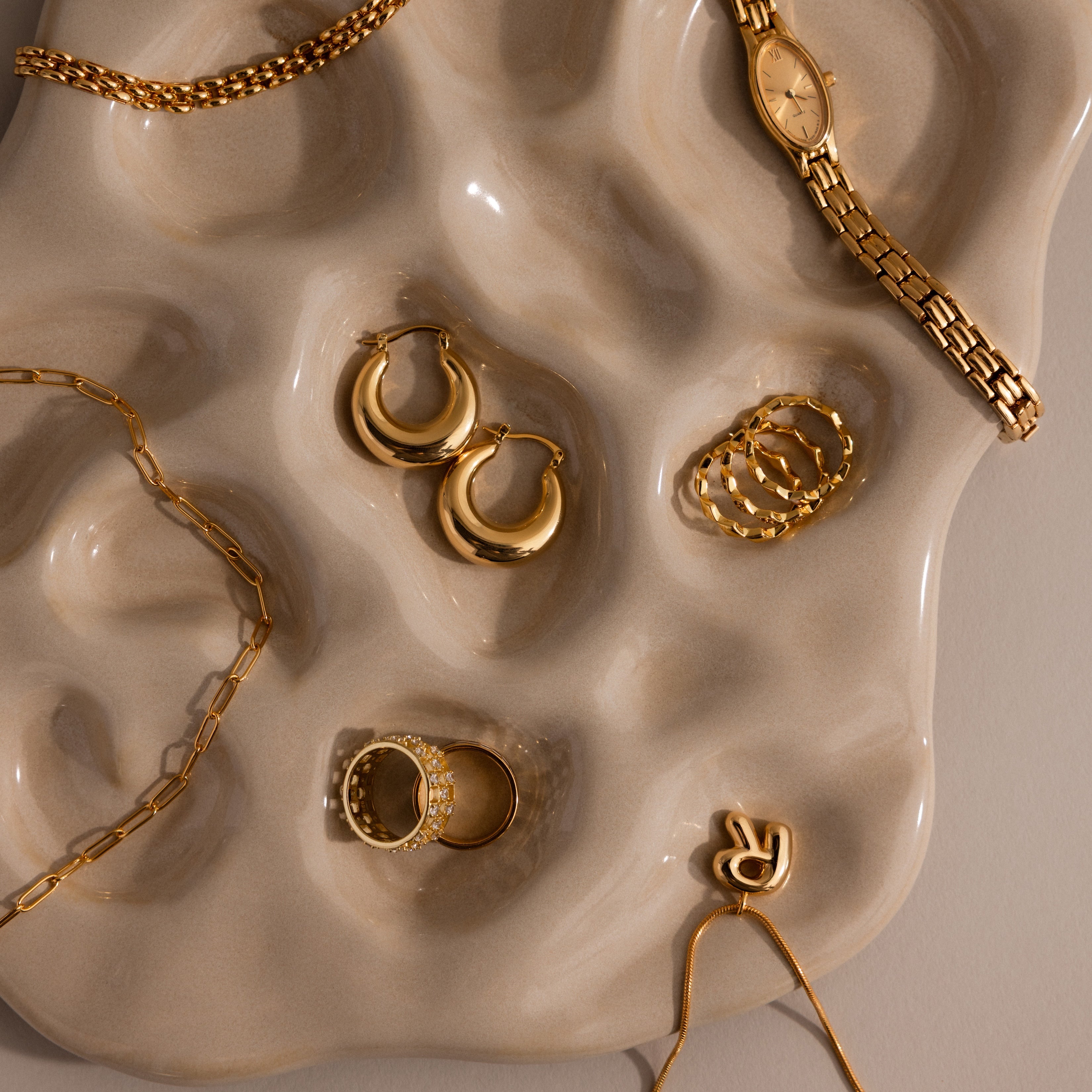 Gold rings, earrings, a bracelet, and a watch displayed on the Organic Treasure Trove Jewelry Dish with a beige textured minimalist design.