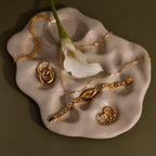 Gold jewelry, the Slim Oval Timepiece with a polished link bracelet, and a white calla lily are arranged on an abstract beige ceramic tray against an olive background.