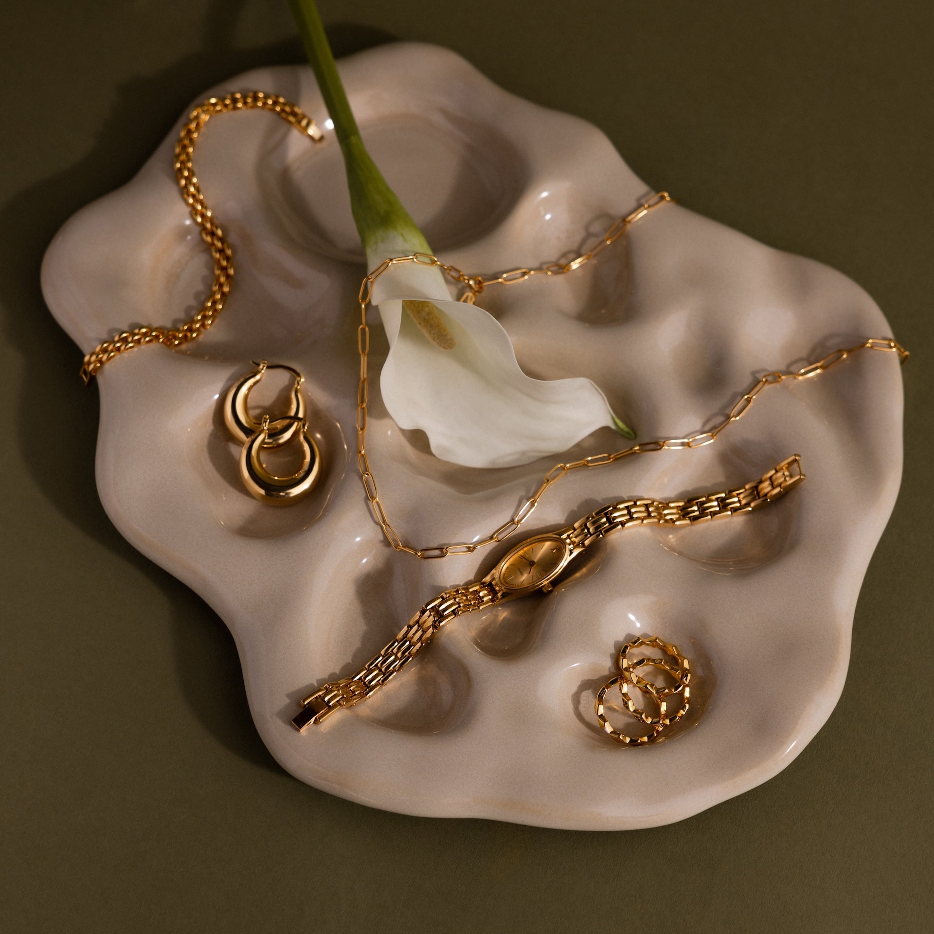 Gold jewelry, the Slim Oval Timepiece with a polished link bracelet, and a white calla lily are arranged on an abstract beige ceramic tray against an olive background.