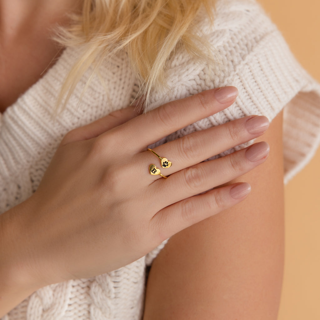 A hand with nude nails wearing a gold Paw Print Duo Heart Ring rests on a white knit sweater—the perfect gift for pet lovers.