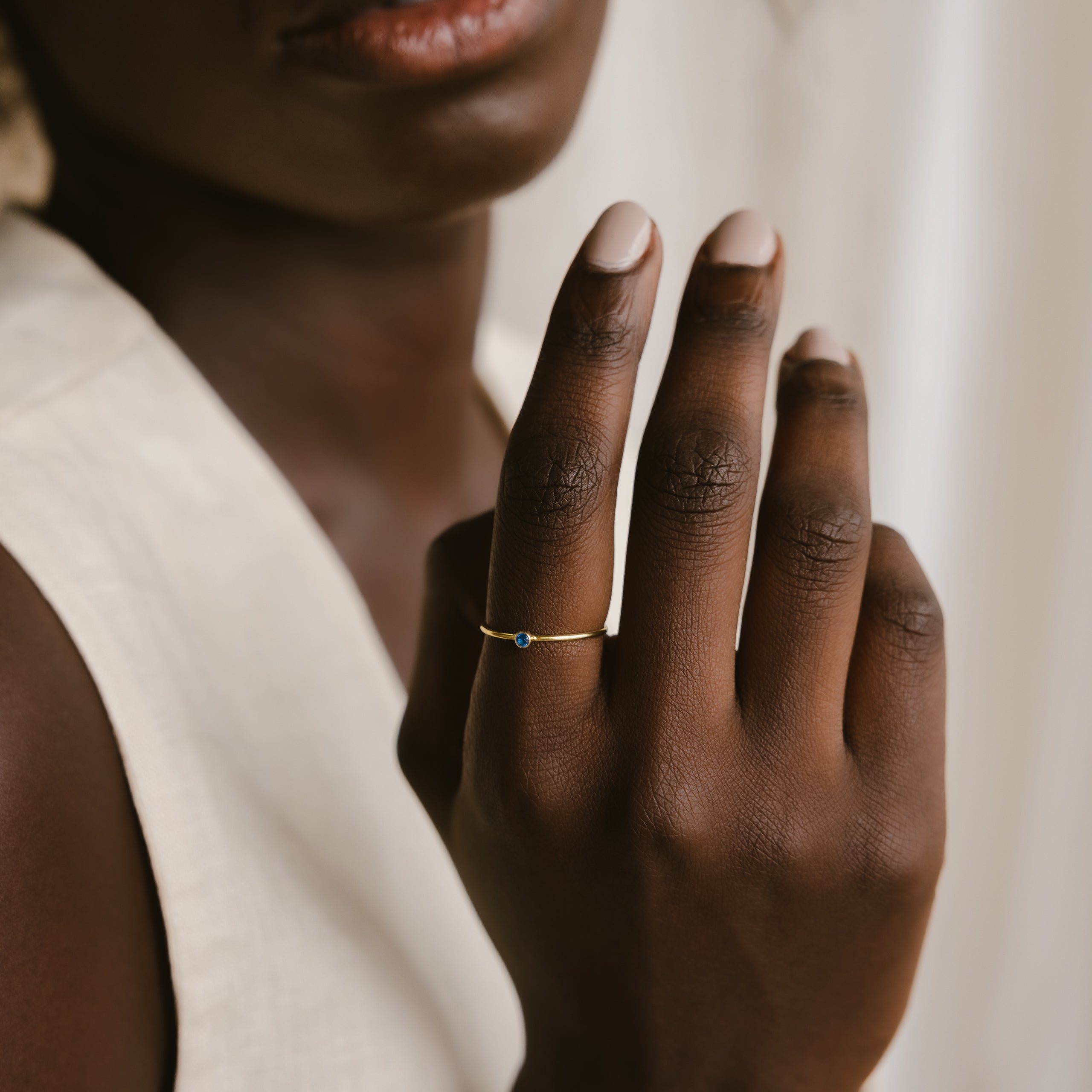 A hand models the Tiny Birthstone Ring with a small blue stone near a woman’s face, highlighting its delicate gemstone detailing against her cream top.