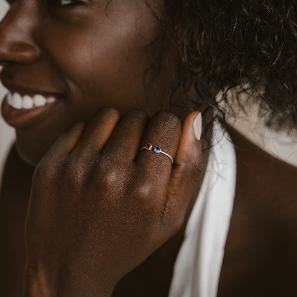 A smiling woman wears the Dual Birthstone Ring, a dainty wrap ring featuring red and blue stones, with her hand resting on her chin.