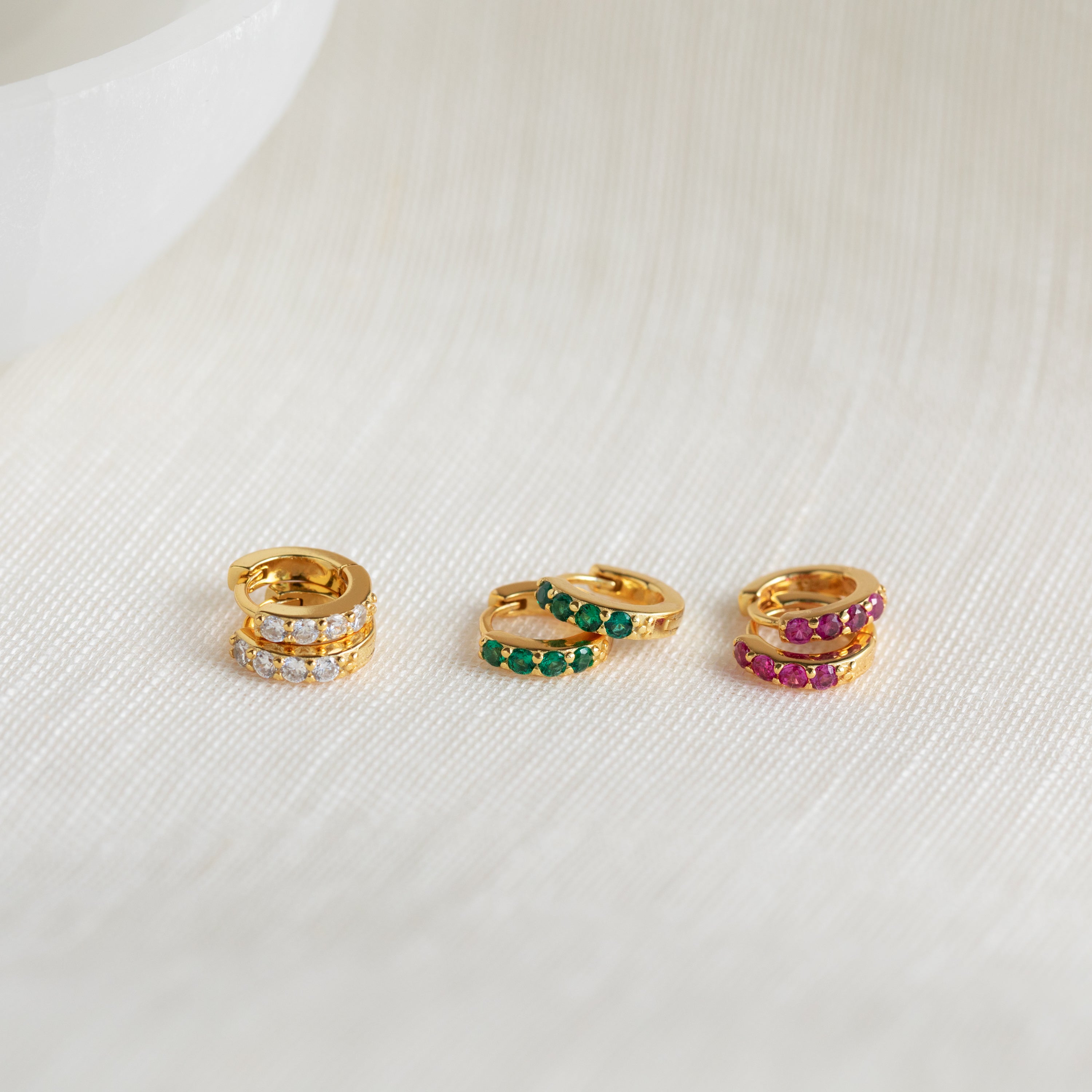 The Elise Gemstone Huggies Set includes three pairs of artisan-made small gold earrings, each featuring white, green, and pink stones displayed on a light textured surface.