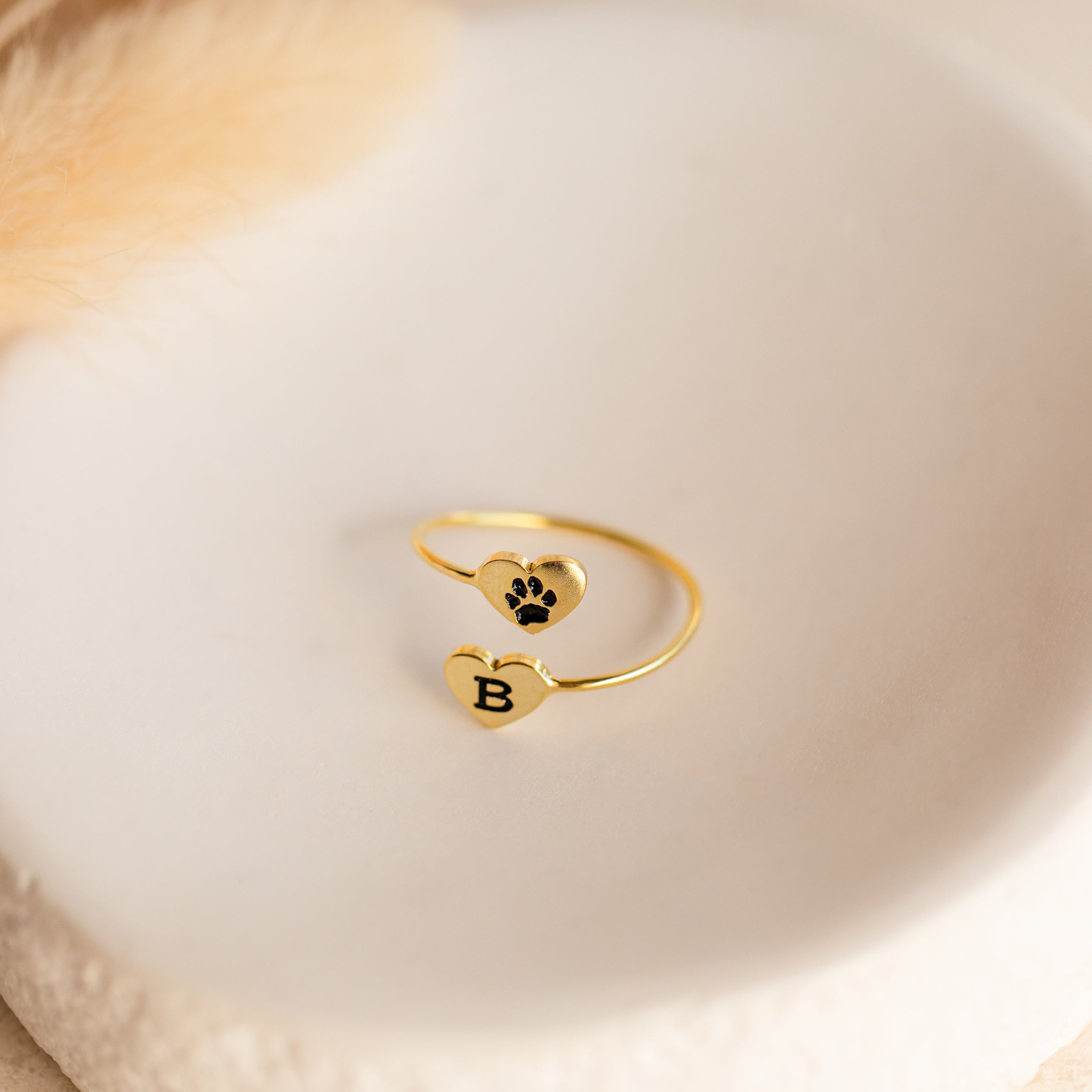 The Paw Print Duo Heart Ring features two gold hearts—one with a black paw print and the other with the letter B—shown on a white dish. A charming Pet Lovers Gift, perfect for showcasing your love for pets.
