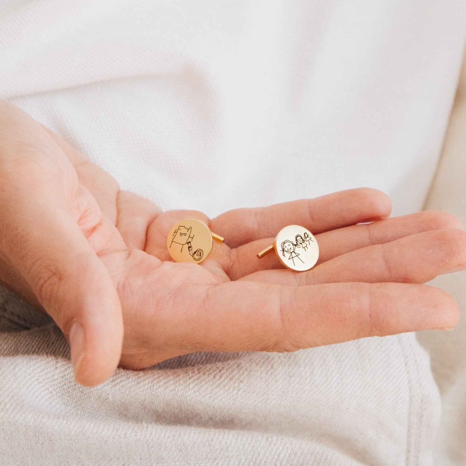 A hand holds two Engraved Cufflinks, each personalized with simple stick-figure designs.