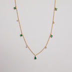 The Larissa Emerald Station Necklace features small green and clear gemstone charms spaced along a gold chain, offering a dainty, vintage-inspired look. Shown on a white background.